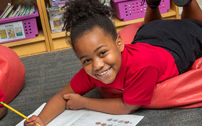 Student smiles at the camera while working in a book on the floor.