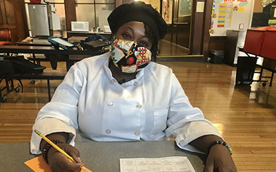 Chef Ronni sits at a table in Baldwin Hall wearing her chef coat and a mask. She has a pencil in her hand and is writing the menu for the week.