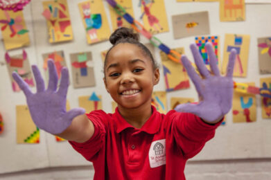 A smiling student who is extending their paint-covered hands towards the camera.