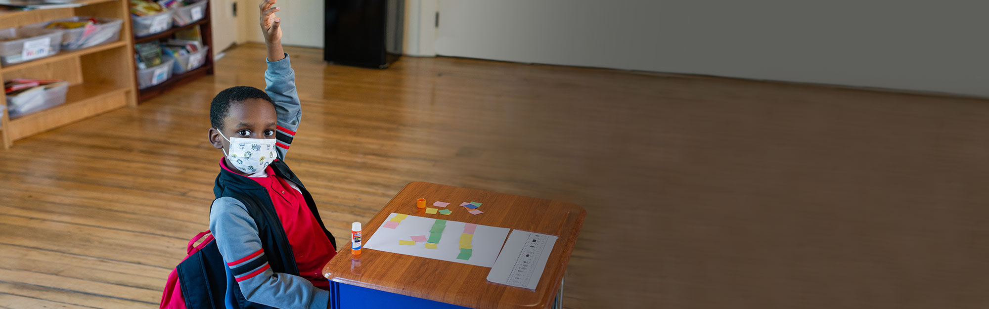 A student sitting at his desk wearing a mask, and raising his hand while looking directly at the camera. He has a glue stick and paper on his desk and is working on a project.