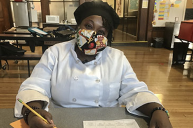 Chef Ronni, wearing a mask and sitting at a table in Balwin Hall. She's holding a pencil and working on the week's menu.