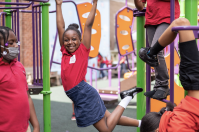 A student jumping into the air and smiling while surrounded by other students on a playground.