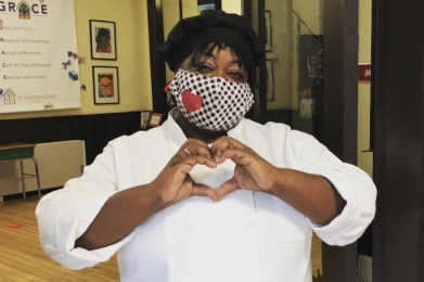 Chef Ronni stands in a doorway wearing a black and white checkered mask with a sparkly heart on it. She is holding her hands in front of her in the shape of a heart.