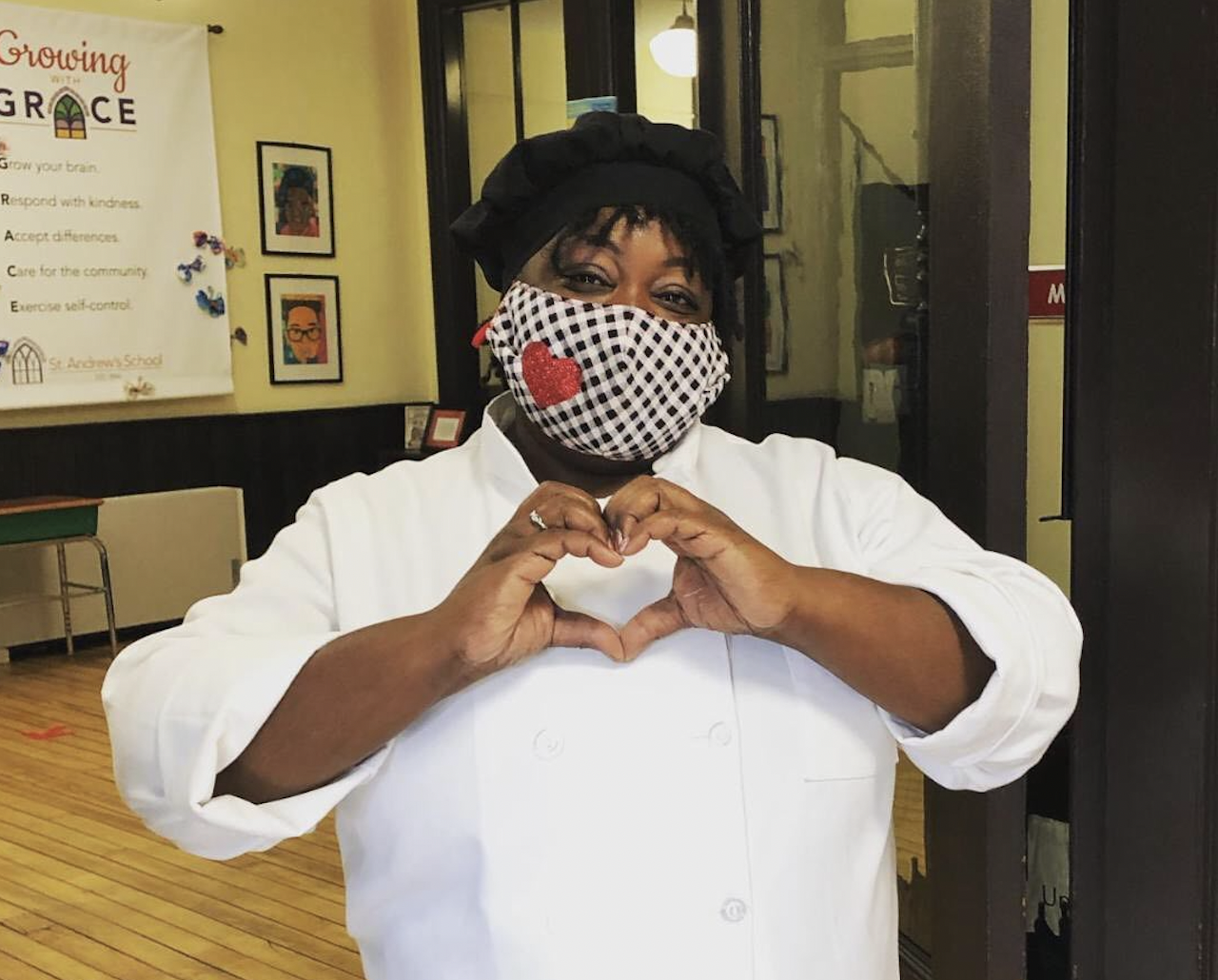 Chef Ronni stands in a doorway wearing a black and white checkered mask with a sparkly heart on it. She is holding her hands in front of her in the shape of a heart.